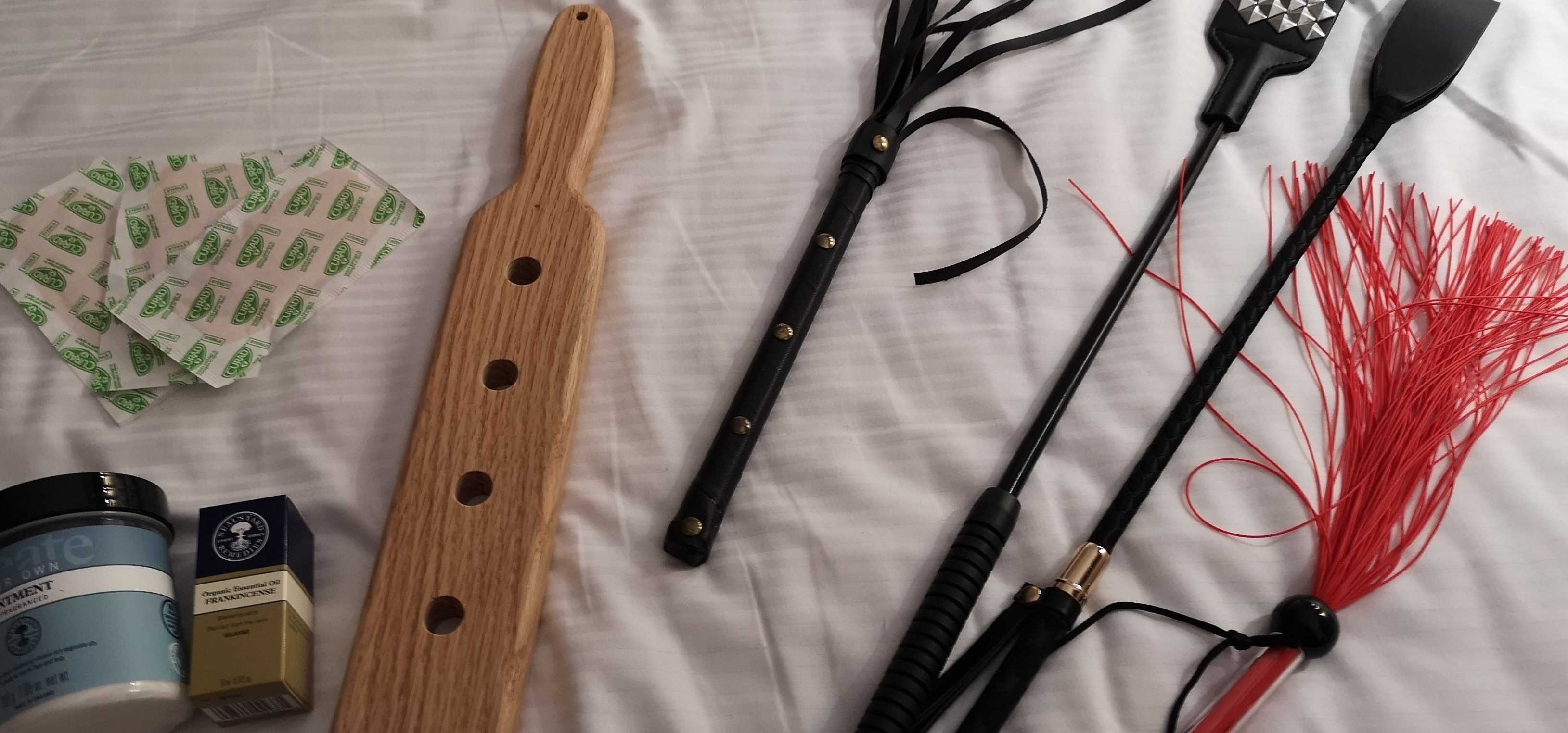 A wooden spanking paddle, a red flogger, a riding crop and a studded riding crop sittng next to some oils and creams